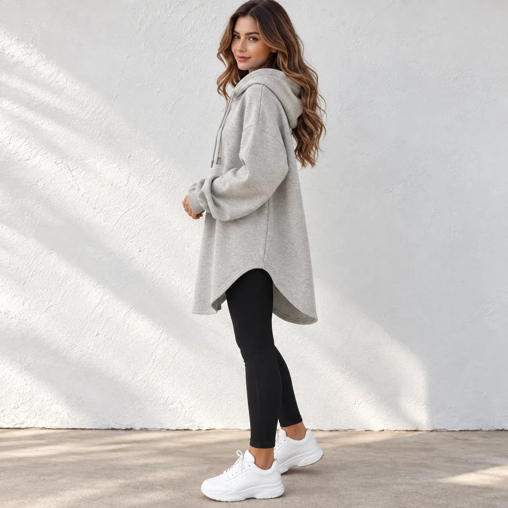 A woman wearing a light gray oversized pullover hoodie with drawstrings, black leggings, and white sneakers, standing against a white textured wall.
