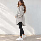 A woman wearing a light gray oversized pullover hoodie with drawstrings, black leggings, and white sneakers, standing against a white textured wall.