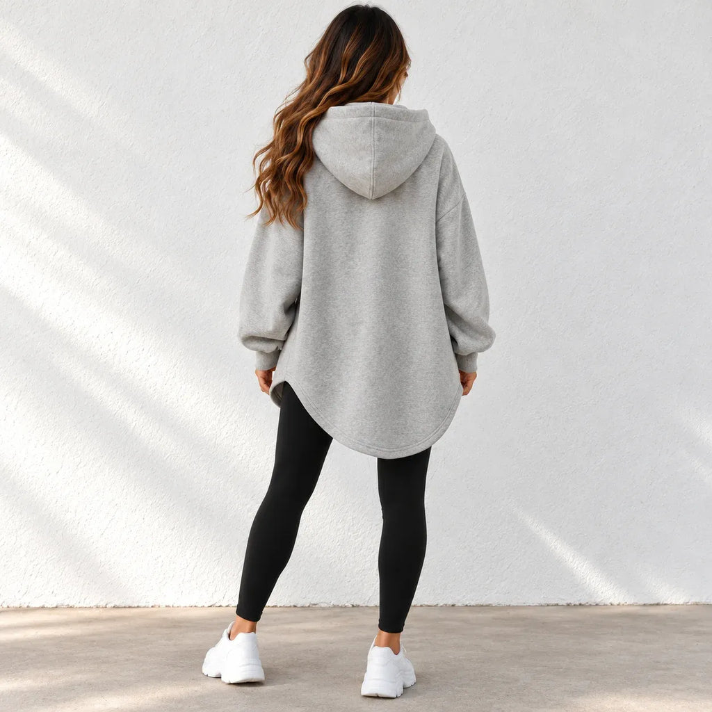 A woman shown from behind wearing a light gray oversized pullover hoodie with black leggings and white sneakers, standing against a white textured wall.