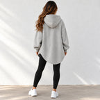 A woman shown from behind wearing a light gray oversized pullover hoodie with black leggings and white sneakers, standing against a white textured wall.