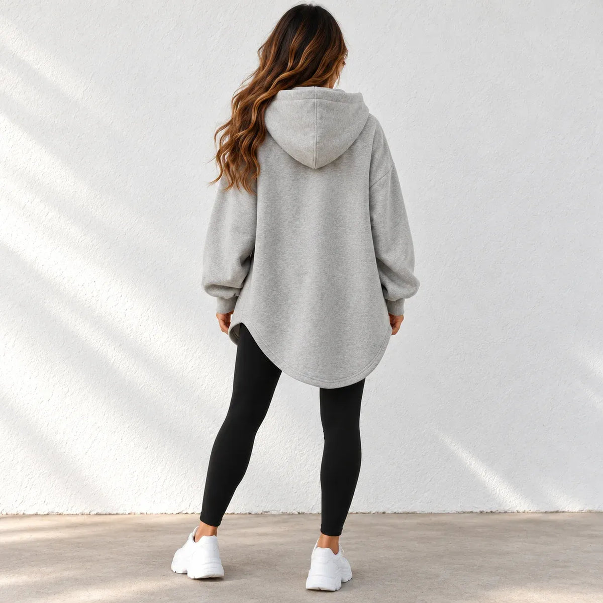 A woman shown from behind wearing a light gray oversized pullover hoodie with black leggings and white sneakers, standing against a white textured wall.