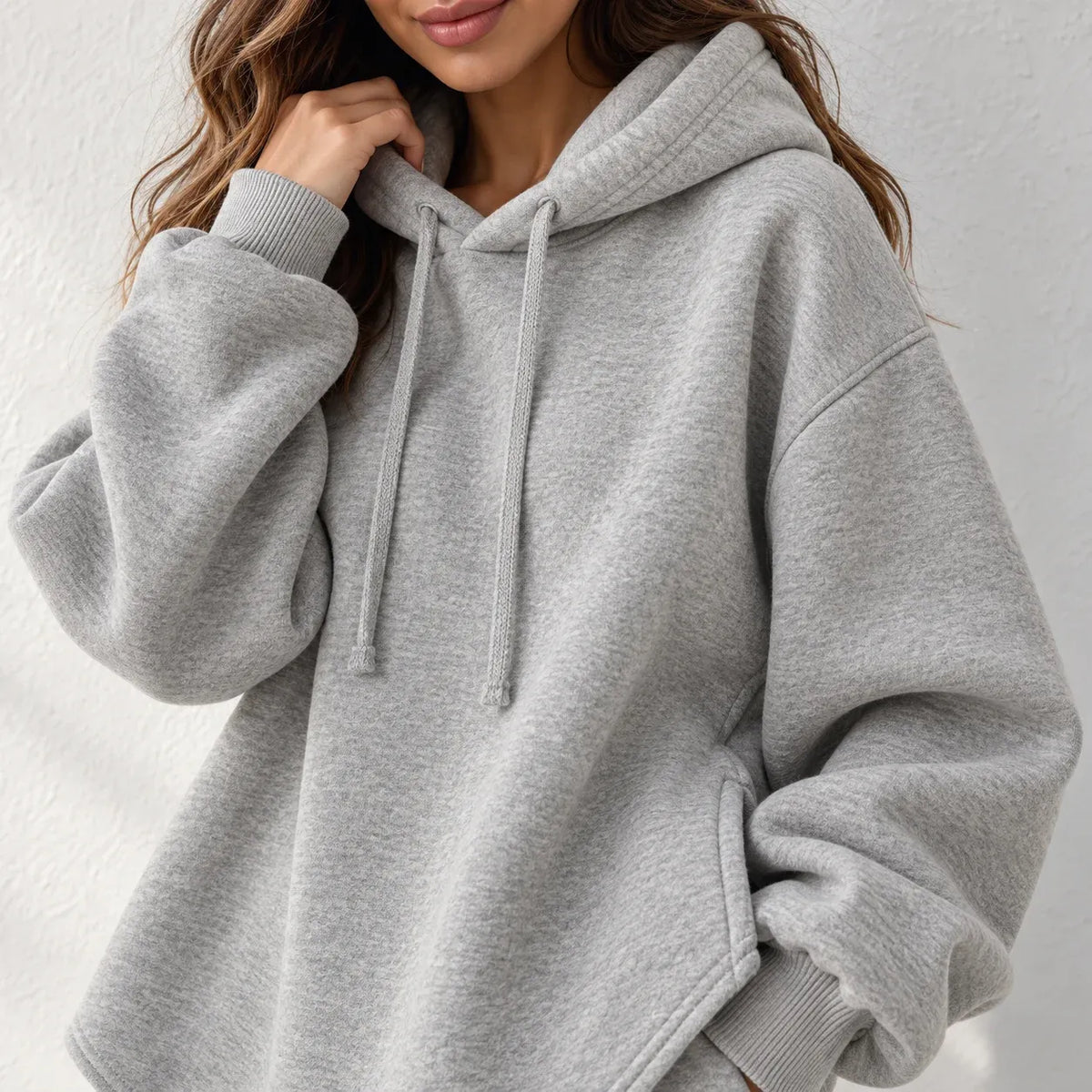 A woman wearing an oversized light gray heather hoodie with ribbed cuffs and drawstrings, shown from the chest up against a plain light wall.