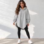 A woman modeling an oversized light gray heathered pullover hoodie with a drawstring hood, paired with black leggings and white sneakers, standing against a white textured wall with soft shadow patterns.