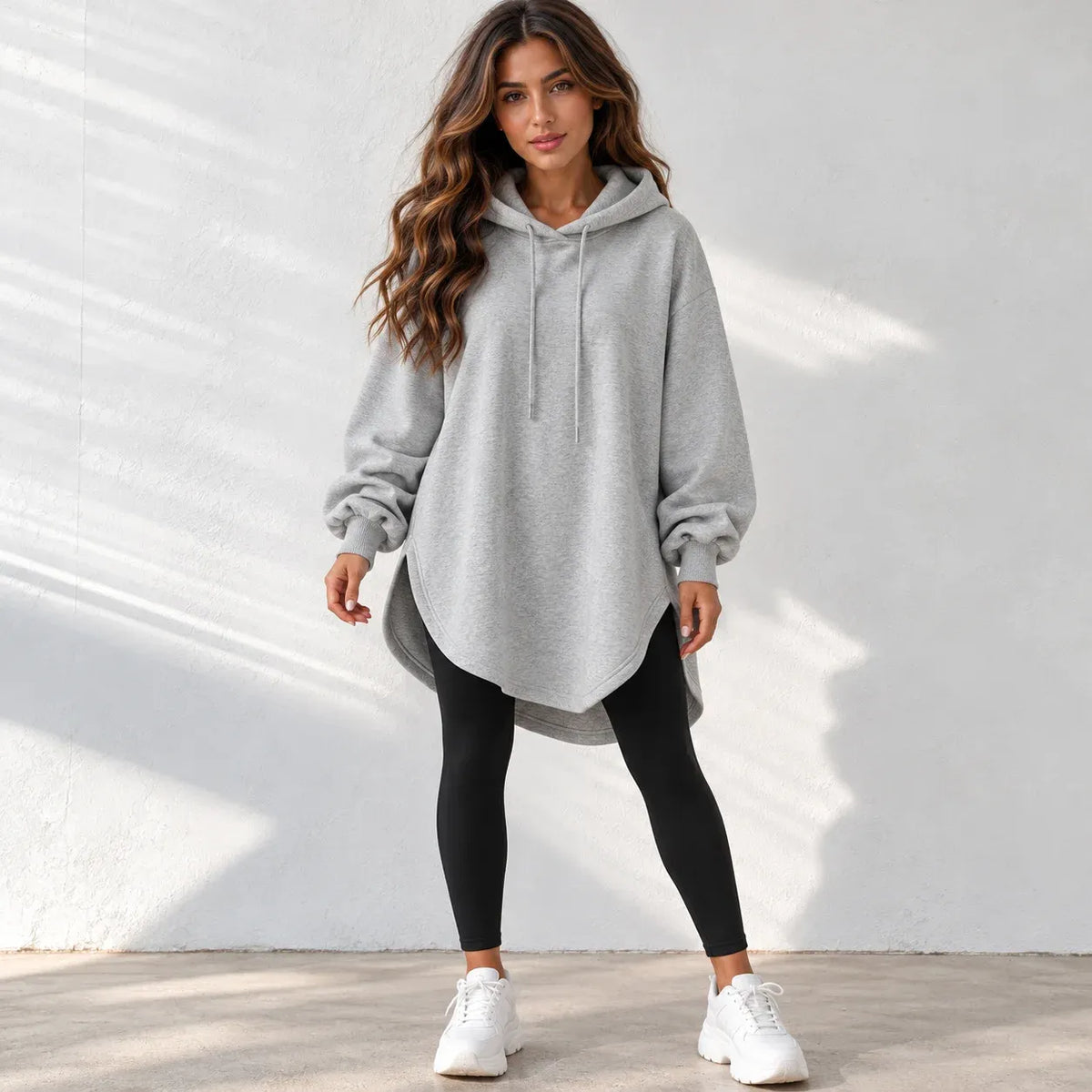 A woman modeling an oversized light gray heathered pullover hoodie with a drawstring hood, paired with black leggings and white sneakers, standing against a white textured wall with soft shadow patterns.