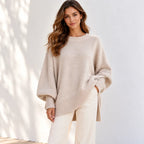 A woman wearing a light beige oversized knit sweater with a crew neck and ribbed trim, paired with white pants, standing against a white wall with soft shadow patterns.