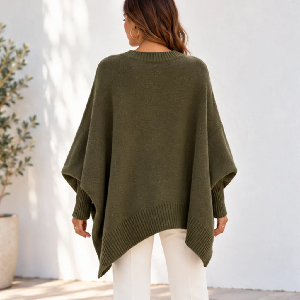 A woman viewed from behind wearing an olive green knit poncho sweater with white pants, standing against a white wall.