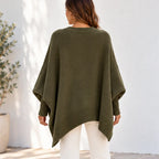 A woman viewed from behind wearing an olive green knit poncho sweater with white pants, standing against a white wall.