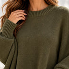 A close-up of a woman wearing an olive green knit sweater, showing the ribbed crew neck, textured knit fabric, and one hand resting near her hair.