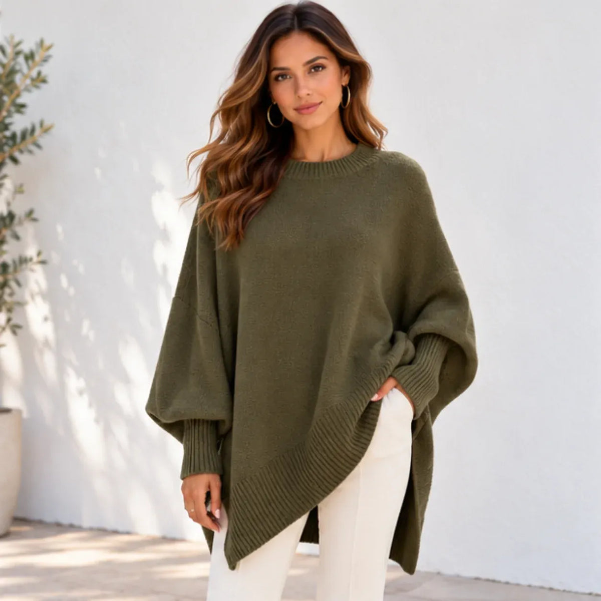 A woman wearing an olive green knit sweater with a relaxed, oversized fit and wide batwing sleeves, paired with white pants, standing against a white stucco wall.