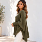 A woman wearing an olive green knit poncho sweater and white pants, standing against a white stucco wall with soft shadows.
