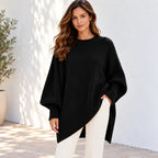 A woman wearing a black oversized knit sweater with wide draped sleeves and an asymmetrical hem, paired with white pants, standing against a white wall with soft shadows.