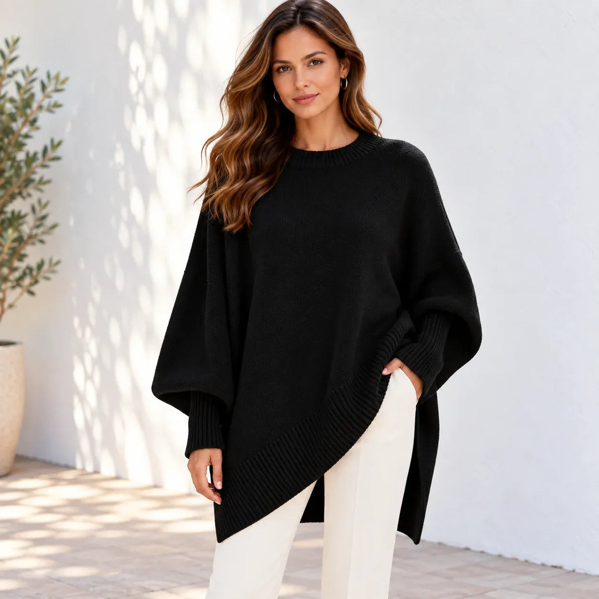 A woman wearing a black oversized knit sweater with wide draped sleeves and an asymmetrical hem, paired with white pants, standing against a white wall with soft shadows.