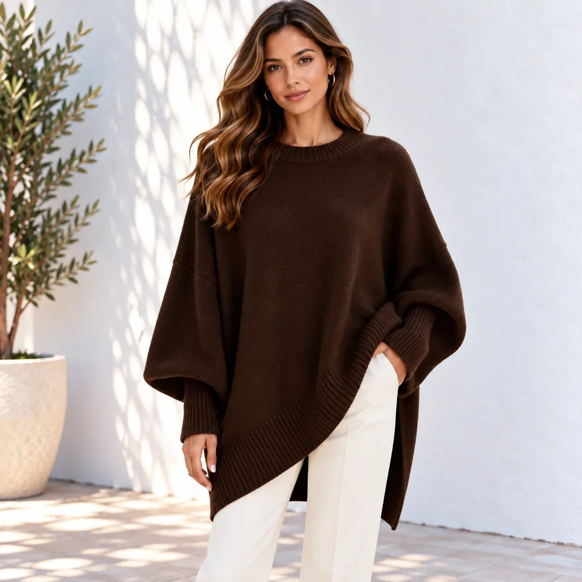 A woman wearing a dark brown oversized knit sweater with wide draped sleeves and a ribbed hem, paired with white pants, standing against a white wall with soft shadows.