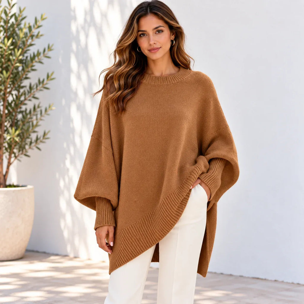 A woman wearing a camel brown knit poncho sweater and white pants, standing against a white wall with a potted plant on the left.