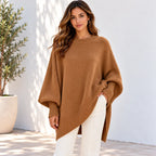 A woman wearing a camel brown knit poncho sweater and white pants, standing against a white wall with a potted plant on the left.
