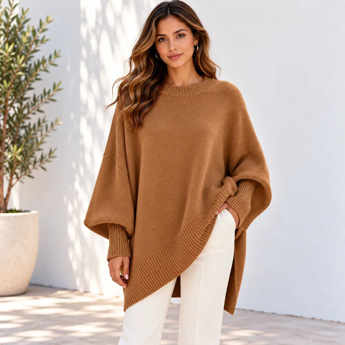 A woman wearing a camel brown knit poncho sweater and white pants, standing against a white wall with a potted plant on the left.