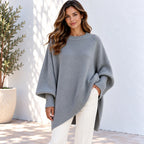 A woman wearing a light gray oversized knit sweater with wide draped sleeves and a ribbed hem, paired with white pants, standing against a white wall with a potted plant on the left.
