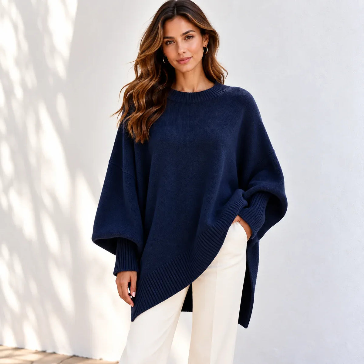 A woman wearing a navy blue knit sweater with a relaxed, oversized fit and white pants, standing against a white wall with soft shadow patterns.