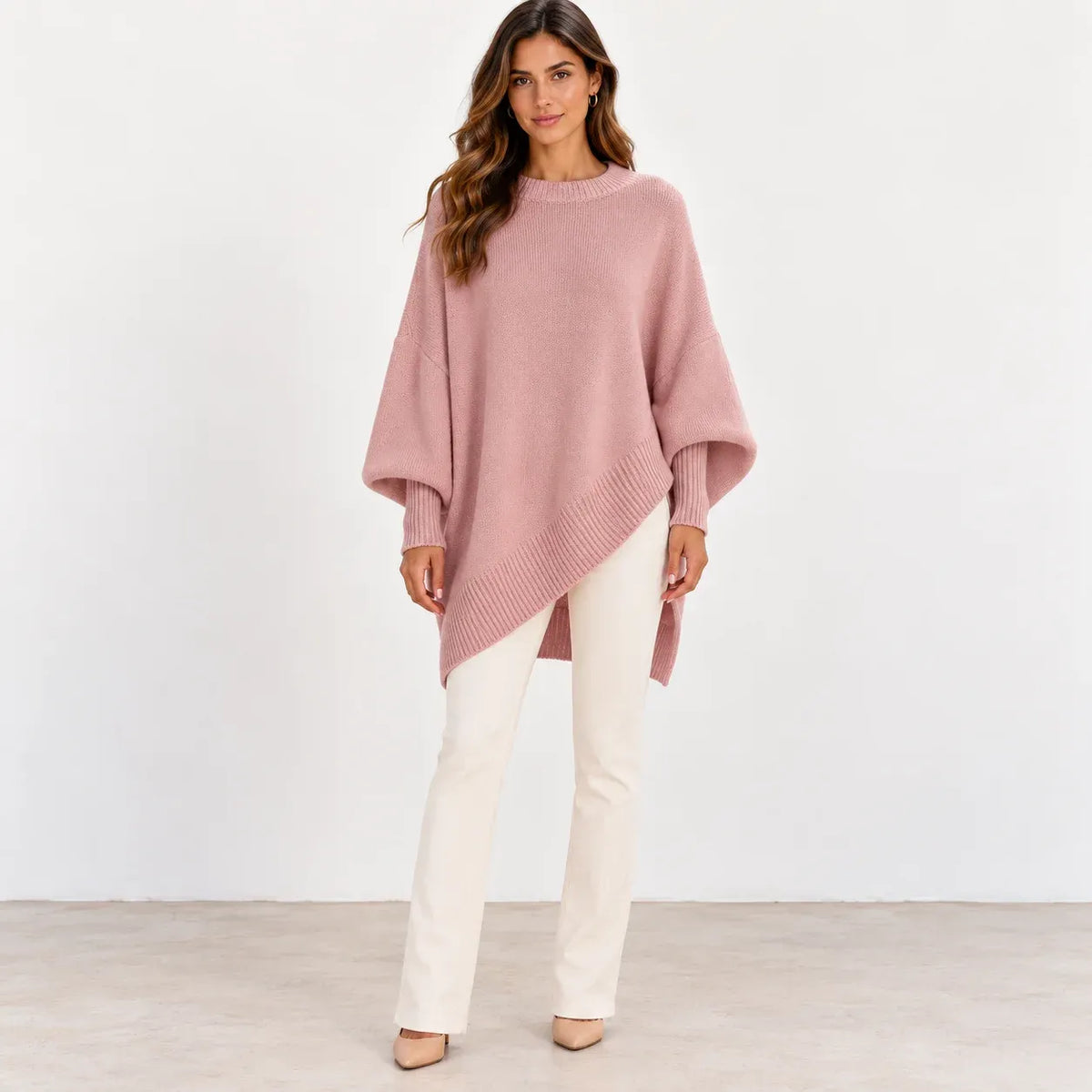 A woman modeling a dusty pink knit poncho sweater with a diagonal hem, paired with off-white pants, standing against a plain white studio wall.