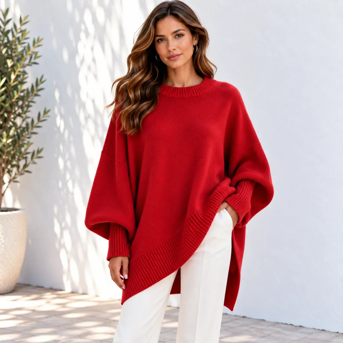 A woman wearing a bright red oversized knit sweater with wide batwing sleeves and an asymmetrical hem, paired with white pants, standing against a white wall with a potted plant on the left.