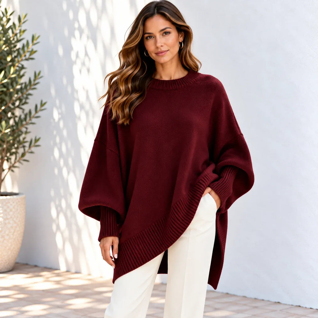 A woman wearing a loose-fitting, oversized burgundy knit sweater with wide batwing sleeves and a ribbed hem, paired with white pants, standing against a white wall with soft shadows.