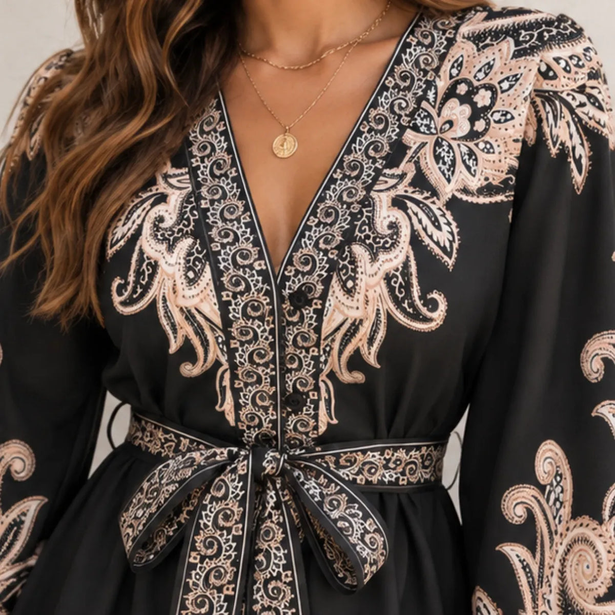 A close-up of a black long-sleeve dress with intricate beige and white paisley embroidery, featuring a V-neckline with a tie belt at the waist.
