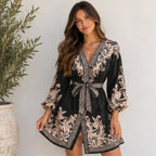 A woman models a black long-sleeve wrap robe with beige and black baroque-style embroidery, featuring a tie waist belt, set against a neutral wall with a potted plant on the left.