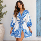 A woman models a white and blue patterned kimono-style cover-up dress with a tie waist belt, standing against a white stucco wall next to a potted plant.
