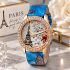 A women's fashion watch with a blue faux-leather strap featuring Paris-themed illustrations, a gold-tone case encrusted with clear rhinestones, and a white dial with Eiffel Tower graphics and red hearts.