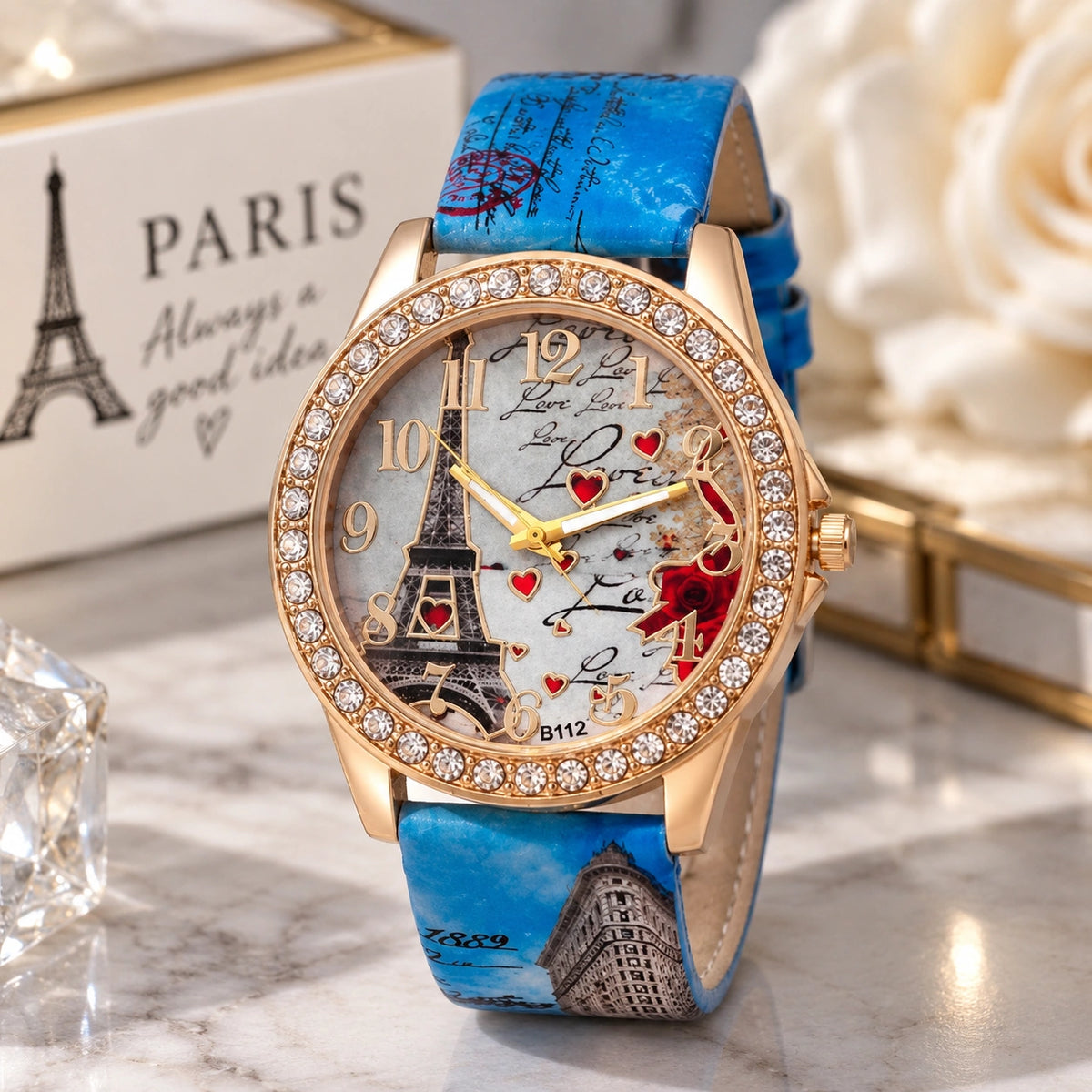 A women's fashion watch with a blue faux-leather strap featuring Paris-themed illustrations, a gold-tone case encrusted with clear rhinestones, and a white dial with Eiffel Tower graphics and red hearts.