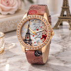 A women's fashion watch with a rose gold-tone case, crystal-studded bezel, and a Paris-themed dial featuring the Eiffel Tower, script writing, and red hearts. The watch has a brown faux-leather strap with a matching Paris print, displayed on a marble surface with decorative props.
