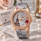 A women's fashion watch with a rose gold case, crystal-studded bezel, and a Paris-themed dial featuring the Eiffel Tower, cursive writing, and red hearts. The watch has a gray distressed leather strap with black script print.