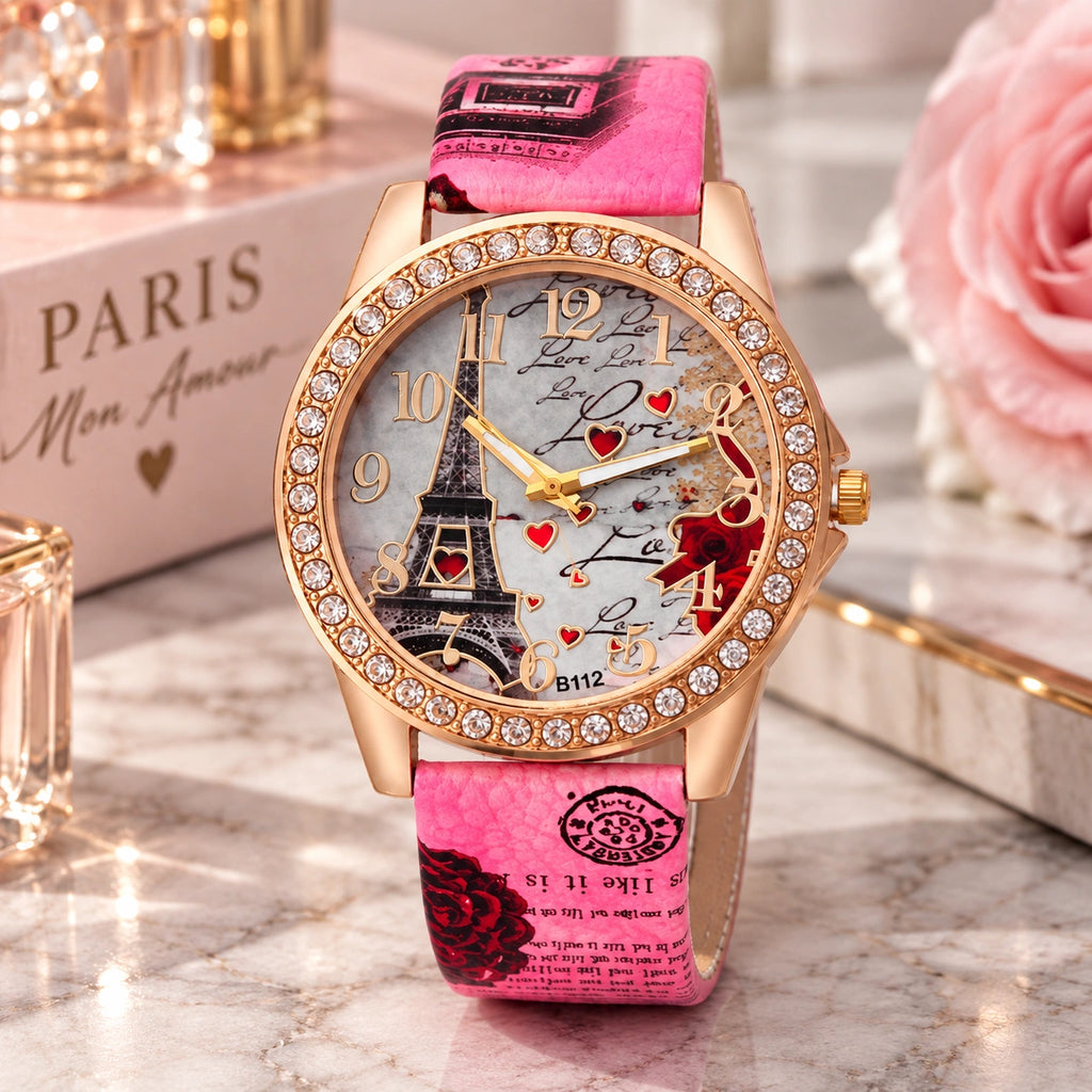 A women's Paris-themed watch with a rose gold-tone case, crystal-studded bezel, and a pink strap printed with Eiffel Tower and Paris motifs. The watch is displayed on a marble surface with decorative props including a pink rose, a book labeled PARIS, and small glass bottles.