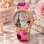 A women's Paris-themed watch with a rose gold-tone case, crystal-studded bezel, and a pink strap printed with Eiffel Tower and Paris motifs. The watch is displayed on a marble surface with decorative props including a pink rose, a book labeled PARIS, and small glass bottles.