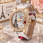 A women's gold-tone watch with a crystal-studded bezel, a white dial featuring an Eiffel Tower design, and a white patterned strap with red rose motifs, displayed on a marble surface with decorative props.