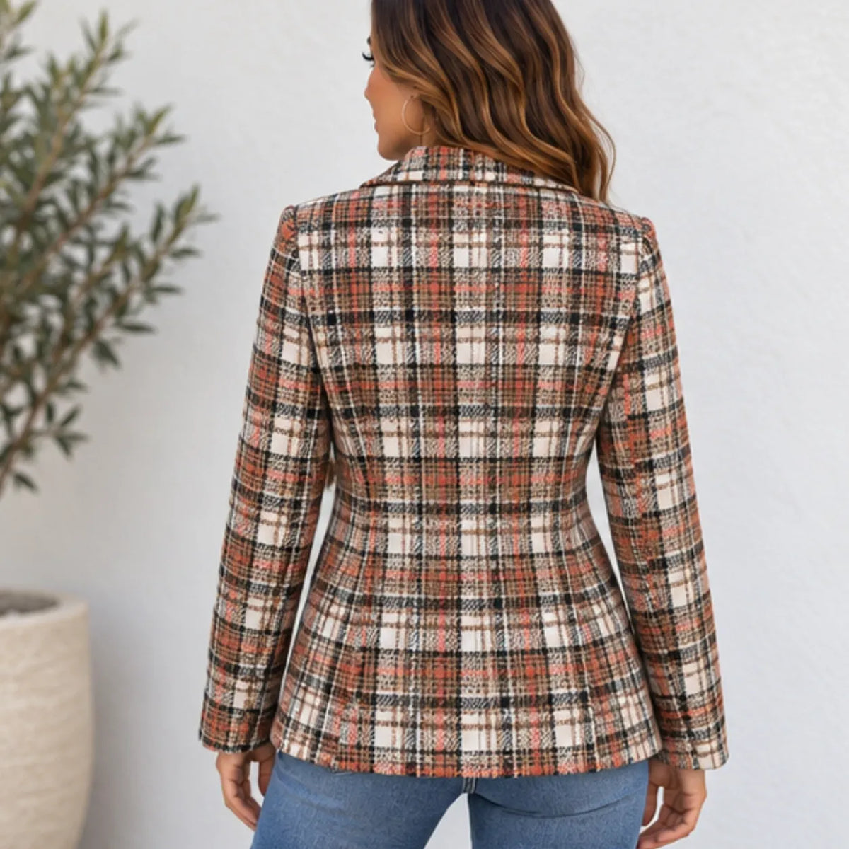 A woman shown from the back wearing a fitted plaid blazer in rust orange, black, and cream, paired with blue jeans, standing against a light gray wall.