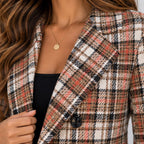 A close-up of a woman wearing a tailored plaid blazer in beige, black, white, and muted red tones over a black ribbed top, with a delicate gold pendant necklace visible.