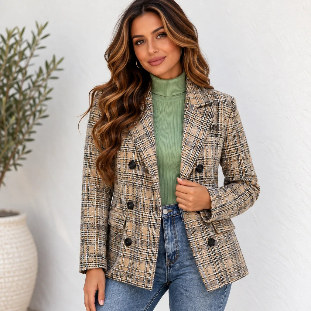 A woman wearing a beige and brown plaid double-breasted blazer over a sage green ribbed turtleneck sweater and blue jeans, standing against a white textured wall.