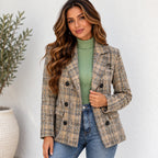 A woman wearing a beige and brown plaid double-breasted blazer over a sage green ribbed turtleneck sweater and blue jeans, standing against a white textured wall.