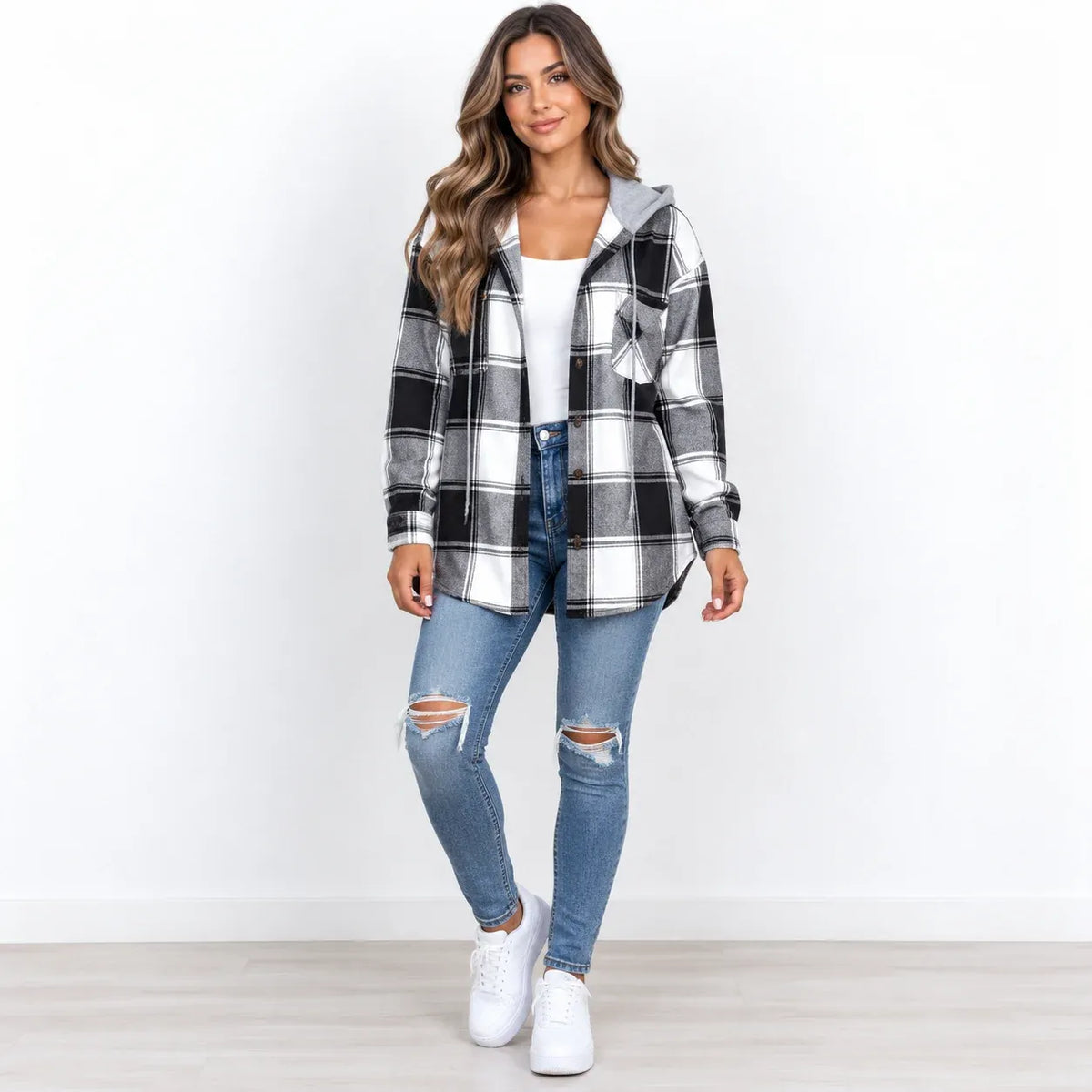A woman modeling a black, white, and gray plaid hooded flannel shirt jacket over a white top and blue ripped jeans, standing against a plain white wall.