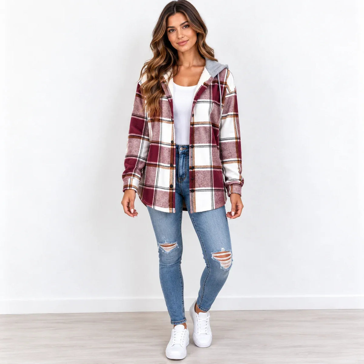 A woman modeling a long-sleeve hooded plaid shirt jacket in burgundy, cream, and gray, worn over a white t-shirt and blue ripped jeans with white sneakers.