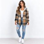 A woman modeling a brown and tan plaid hooded button-up shirt jacket over a white top, paired with distressed blue jeans and white sneakers, standing against a white studio background.