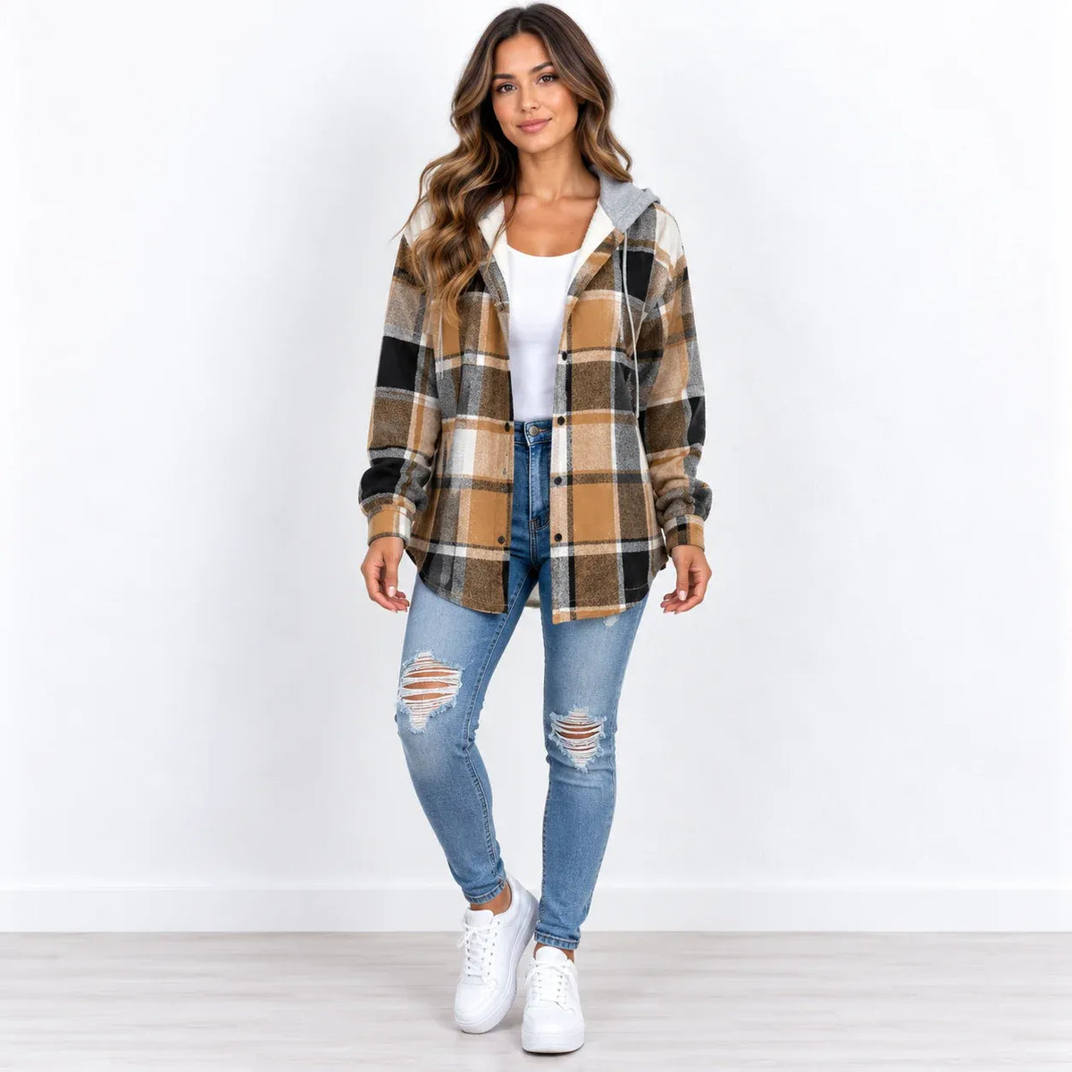 A woman modeling a brown and tan plaid hooded button-up shirt jacket over a white top, paired with distressed blue jeans and white sneakers, standing against a white studio background.