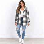 A woman modeling a gray, white, and black plaid hooded button-up shirt jacket, paired with a white top, distressed blue jeans, and white sneakers, set against a plain white wall and light wood floor.