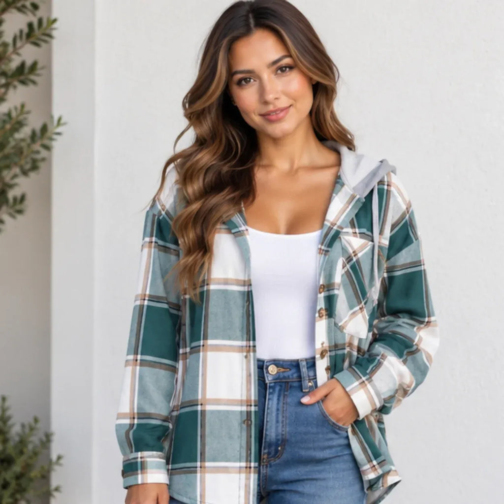 A woman wearing a green, white, and brown plaid hooded button-up shirt over a white tank top and blue jeans, standing against a light-colored wall.