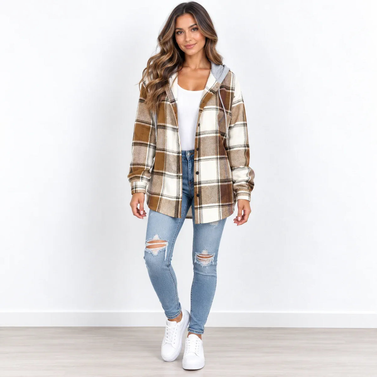 A woman modeling a brown and beige plaid hooded button-up shirt jacket over a white top, paired with light blue distressed jeans and white sneakers, standing against a white wall.
