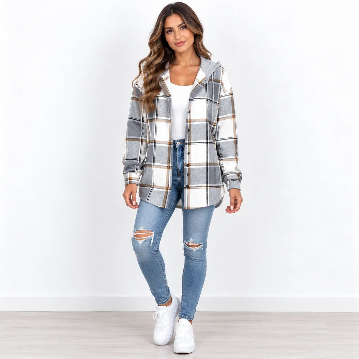 A woman modeling a gray and white plaid hooded button-up shirt jacket over a white top and distressed blue jeans, standing against a plain white wall.