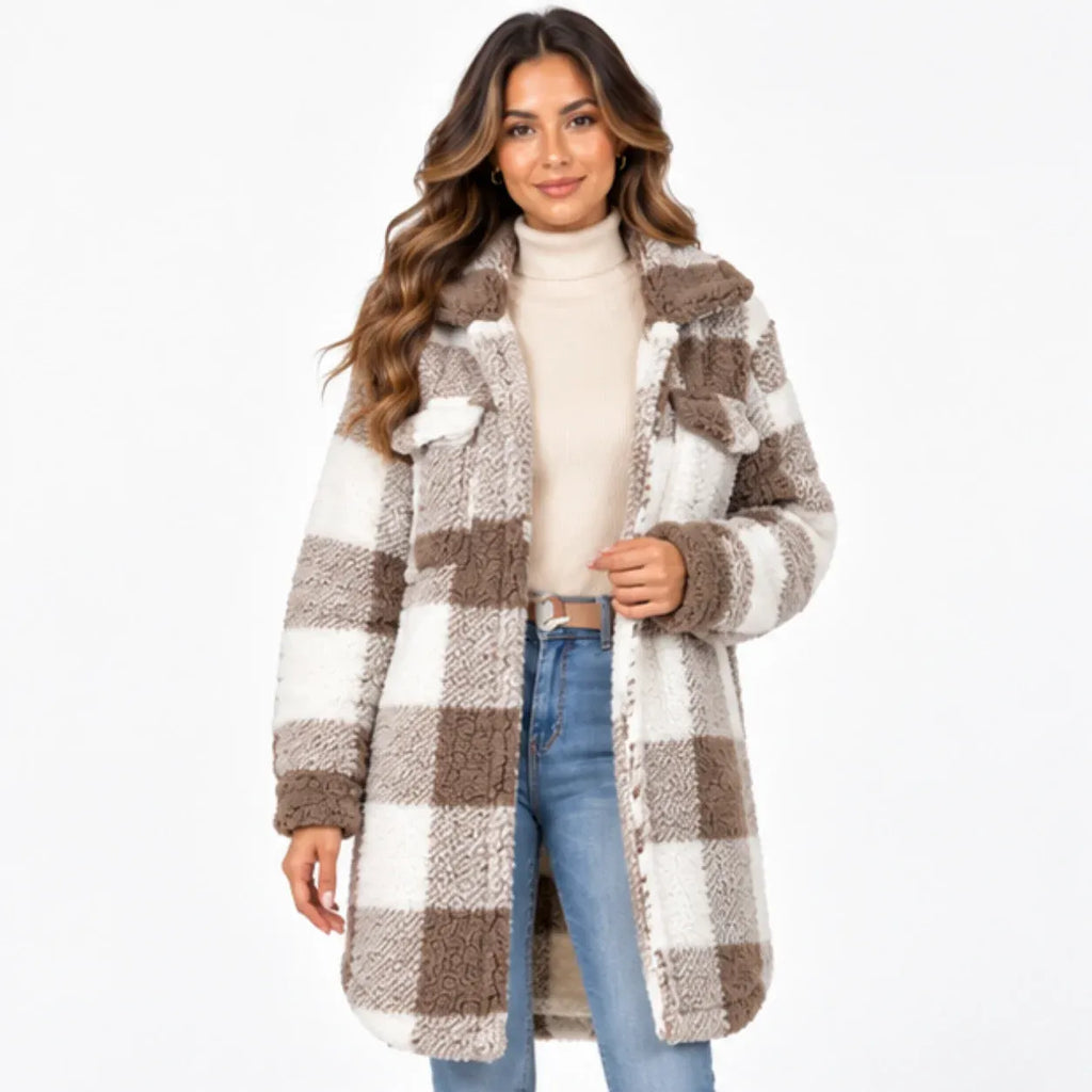 A woman modeling a mid-length, oversized teddy fleece coat with a brown and cream plaid pattern, worn over a cream turtleneck and blue jeans against a plain white background.