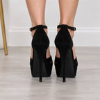 Black high-heeled platform sandals with ankle straps, shown from the back on a model's feet against a light gray floor and white wall.