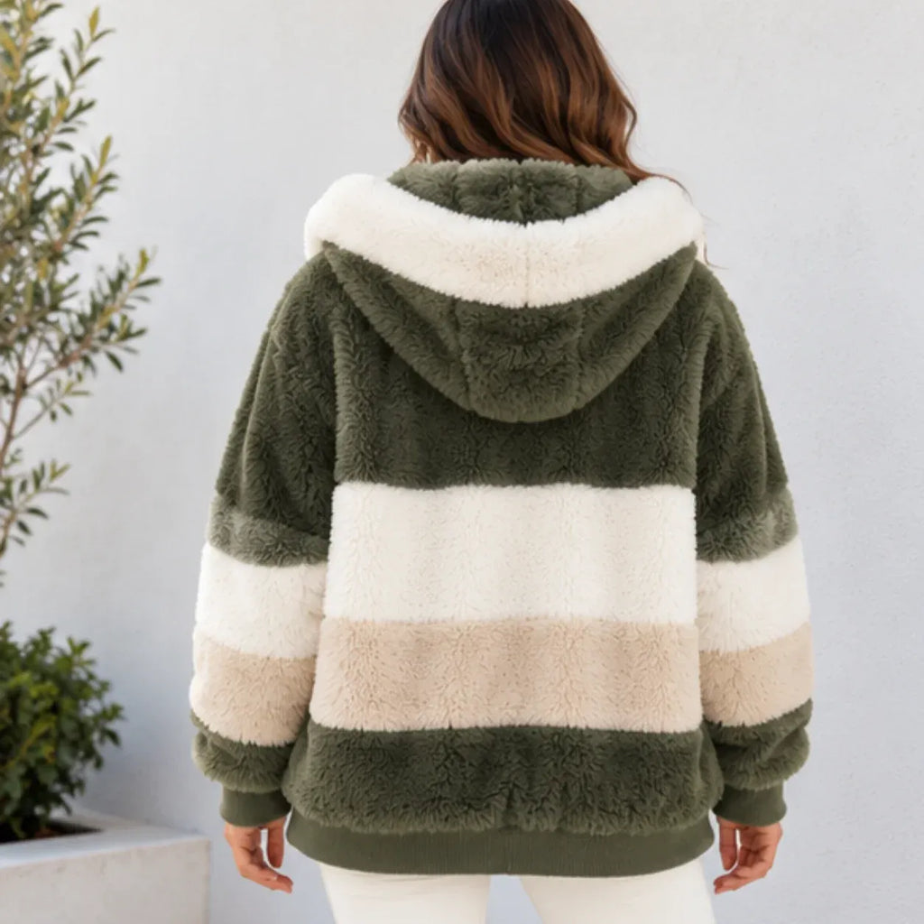 A woman viewed from behind wearing a plush hooded jacket with wide horizontal stripes in olive green, cream, and beige, standing against a light gray wall with a plant at the side.